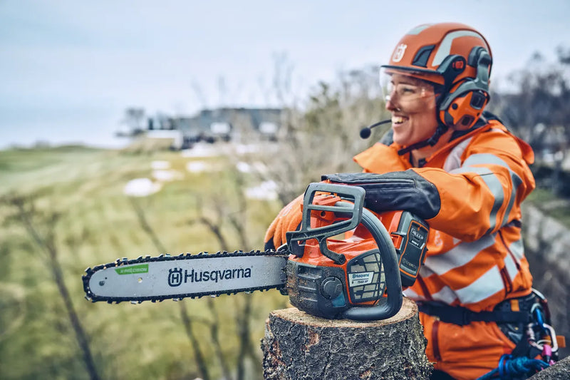 The Husqvarna T542i XP Battery Chainsaw (Skin Only) features an eye-catching orange and black body, a sturdy metal chain bar with the "Husqvarna" brand name displayed on it, a grip handle, and various adjustment controls against a transparent background.