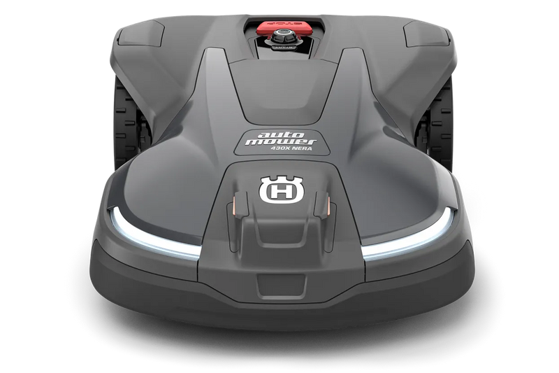 The Husqvarna Automower 430X NERA is a sleek, grey robotic lawn mower featuring two large wheels at the rear and two smaller wheels at the front. It boasts a red stop button on top, LED headlights, and proudly displays the Husqvarna logo on the front. With its modern design, this mower is equipped with object avoidance technology and can be conveniently controlled via the Automower Connect app.