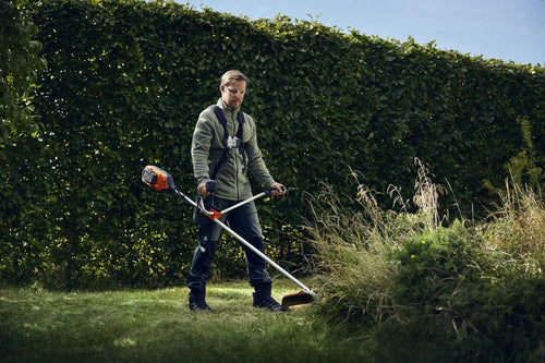 A Husqvarna 325iR Brushcutter (skin) lays on a white background. Perfect for lawns and other outdoor maintenance, the tool features a long silver shaft with a black handle at the top and an orange cutting head at the bottom. The motor is housed in an orange casing near the handle.