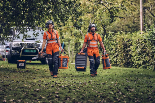 A pair of Husqvarna Arbor Waist trousers, Technical Extreme, in orange and black with attached suspenders designed for climbing and working in trees. These trousers feature multiple pockets, reinforced knee areas, and adjustable straps branded with "Husqvarna." The rugged design offers safety, agility, and functionality for demanding tasks.