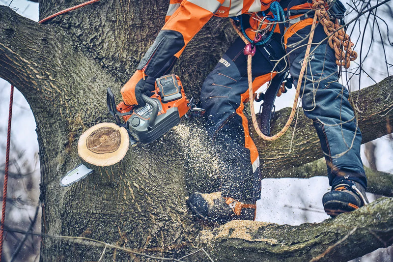 The Husqvarna T542i XP Battery Chainsaw (Skin Only) features an eye-catching orange and black body, a sturdy metal chain bar with the "Husqvarna" brand name displayed on it, a grip handle, and various adjustment controls against a transparent background.