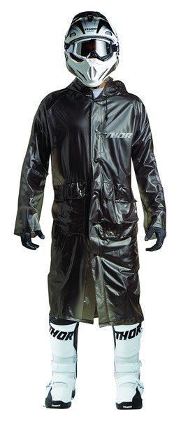 A person sports a comprehensive motocross ensemble, showcasing a TRENCH COAT THOR MX EXCEL RAIN in black with improved ventilation, alongside a helmet, gloves, and white boots stamped with "Thor." This gear is ideal for off-road biking in wet conditions.