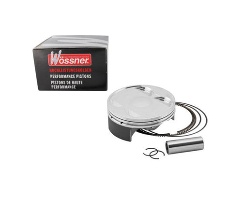 Image of a WOSSNER PISTON KIT WOSSNER KAWASAKI KX250F 04-05 SUZUKI RMZ250 04-06 76.97MM, featuring a piston, piston rings, wrist pin, and circlips, all neatly arranged beside a branded box labeled "HIGH PERFORMANCE PISTONS" in three languages.
