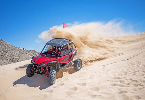 A red Honda Talon R off-road vehicle is pictured. It features robust, large tires, a roll cage, and sporty accents. The vehicle has a rugged design suitable for rough terrain, with visible shock absorbers and a roof for protection—perfect for sport side-by-side enthusiasts.
