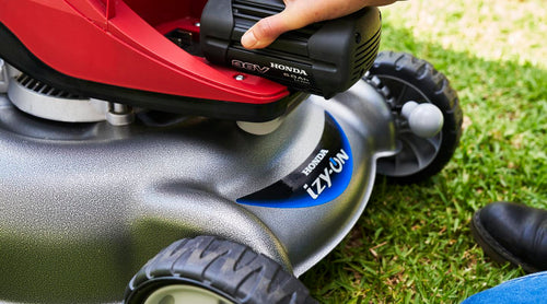 The Honda HRG466 Battery izy-ON Lawnmower Kit by Honda Power Equipment features a silver and black design with a red engine cover that evokes the look of an MX motorbike. It has a black handlebar with a grip, four wheels for stability, and comes equipped with a black grass collection bag at the back. The engine cover displays various warning labels for user safety.