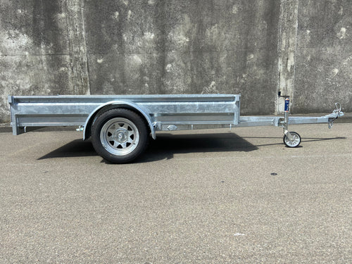 A City Honda Manawatu Custom Built 8x4 Tip Trailer with a single axle, tilted upward at the front where it connects to a vehicle, is parked on a paved road against a concrete wall. The trailer has a silver finish and is in good condition, ideal for transporting bikes or generators.