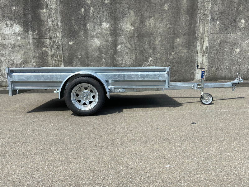A City Honda Manawatu Custom Built 8x4 Tip Trailer with a single axle, tilted upward at the front where it connects to a vehicle, is parked on a paved road against a concrete wall. The trailer has a silver finish and is in good condition, ideal for transporting bikes or generators.
