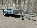 A City Honda Manawatu Custom Built 8x4 Tip Trailer with a single axle, tilted upward at the front where it connects to a vehicle, is parked on a paved road against a concrete wall. The trailer has a silver finish and is in good condition, ideal for transporting bikes or generators.