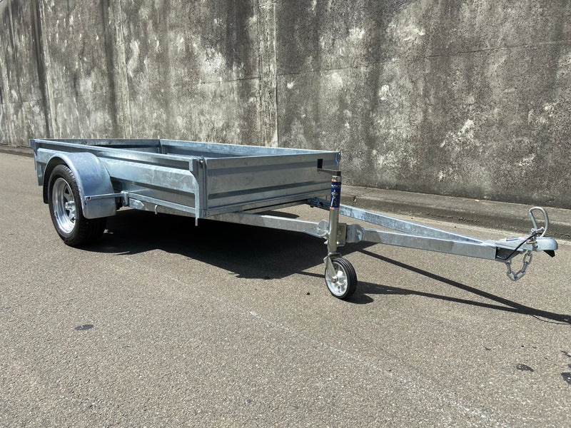 A City Honda Manawatu Custom Built 8x4 Tip Trailer with a single axle, tilted upward at the front where it connects to a vehicle, is parked on a paved road against a concrete wall. The trailer has a silver finish and is in good condition, ideal for transporting bikes or generators.