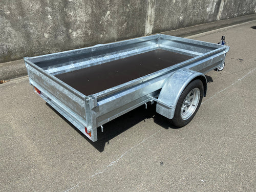 A City Honda Manawatu Custom Built 8x4 Tip Trailer with a single axle, tilted upward at the front where it connects to a vehicle, is parked on a paved road against a concrete wall. The trailer has a silver finish and is in good condition, ideal for transporting bikes or generators.