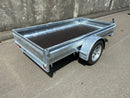 A City Honda Manawatu Custom Built 8x4 Tip Trailer with a single axle, tilted upward at the front where it connects to a vehicle, is parked on a paved road against a concrete wall. The trailer has a silver finish and is in good condition, ideal for transporting bikes or generators.