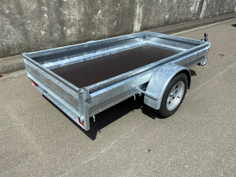 A City Honda Manawatu Custom Built 8x4 Tip Trailer with a single axle, tilted upward at the front where it connects to a vehicle, is parked on a paved road against a concrete wall. The trailer has a silver finish and is in good condition, ideal for transporting bikes or generators.