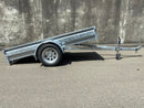 A City Honda Manawatu Custom Built 8x4 Tip Trailer with a single axle, tilted upward at the front where it connects to a vehicle, is parked on a paved road against a concrete wall. The trailer has a silver finish and is in good condition, ideal for transporting bikes or generators.
