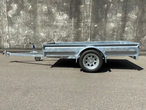 A City Honda Manawatu Custom Built 8x4 Tip Trailer with a single axle, tilted upward at the front where it connects to a vehicle, is parked on a paved road against a concrete wall. The trailer has a silver finish and is in good condition, ideal for transporting bikes or generators.