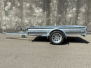 A City Honda Manawatu Custom Built 8x4 Tip Trailer with a single axle, tilted upward at the front where it connects to a vehicle, is parked on a paved road against a concrete wall. The trailer has a silver finish and is in good condition, ideal for transporting bikes or generators.