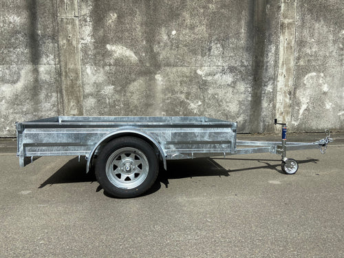 A Custom Built 7x4 Trailer by City Honda Manawatu with a metal frame and a small wheel jack at the front, positioned on a concrete surface against a weathered wall. The trailer has a simple design, ideal for hauling items or equipment from gardens or even motorbikes like your Honda.