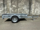 A Custom Built 7x4 Trailer by City Honda Manawatu with a metal frame and a small wheel jack at the front, positioned on a concrete surface against a weathered wall. The trailer has a simple design, ideal for hauling items or equipment from gardens or even motorbikes like your Honda.