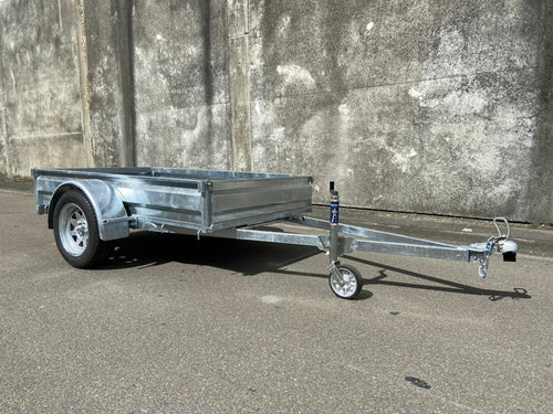 A Custom Built 7x4 Trailer by City Honda Manawatu with a metal frame and a small wheel jack at the front, positioned on a concrete surface against a weathered wall. The trailer has a simple design, ideal for hauling items or equipment from gardens or even motorbikes like your Honda.