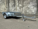 A Custom Built 7x4 Trailer by City Honda Manawatu with a metal frame and a small wheel jack at the front, positioned on a concrete surface against a weathered wall. The trailer has a simple design, ideal for hauling items or equipment from gardens or even motorbikes like your Honda.
