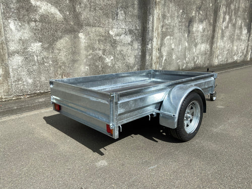 A Custom Built 7x4 Trailer by City Honda Manawatu with a metal frame and a small wheel jack at the front, positioned on a concrete surface against a weathered wall. The trailer has a simple design, ideal for hauling items or equipment from gardens or even motorbikes like your Honda.