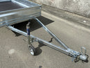 A Custom Built 7x4 Trailer by City Honda Manawatu with a metal frame and a small wheel jack at the front, positioned on a concrete surface against a weathered wall. The trailer has a simple design, ideal for hauling items or equipment from gardens or even motorbikes like your Honda.