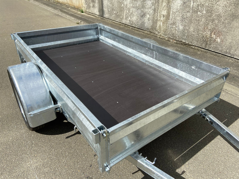 A Custom Built 7x4 Trailer by City Honda Manawatu with a metal frame and a small wheel jack at the front, positioned on a concrete surface against a weathered wall. The trailer has a simple design, ideal for hauling items or equipment from gardens or even motorbikes like your Honda.