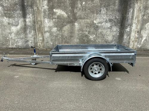 A Custom Built 7x4 Trailer by City Honda Manawatu with a metal frame and a small wheel jack at the front, positioned on a concrete surface against a weathered wall. The trailer has a simple design, ideal for hauling items or equipment from gardens or even motorbikes like your Honda.