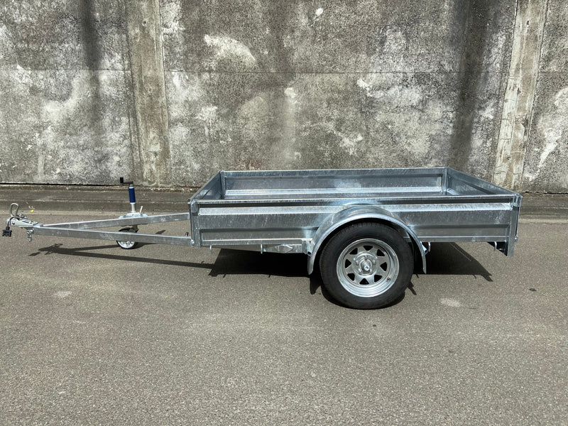 A Custom Built 7x4 Trailer by City Honda Manawatu with a metal frame and a small wheel jack at the front, positioned on a concrete surface against a weathered wall. The trailer has a simple design, ideal for hauling items or equipment from gardens or even motorbikes like your Honda.