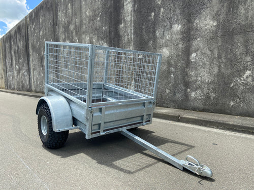 A City Honda Manawatu Custom Built Caged ATV Trailer is parked on a concrete surface. The trailer's tongue is extended and rests on the ground, beside several Honda generators. A weathered concrete wall forms the backdrop.