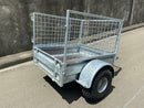 A City Honda Manawatu Custom Built Caged ATV Trailer is parked on a concrete surface. The trailer's tongue is extended and rests on the ground, beside several Honda generators. A weathered concrete wall forms the backdrop.