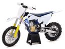 A white and blue dirt bike with a yellow front fork is displayed on a black stand. The bike, a MODEL DIRT BIKE HUSQVARNA FC450 1:12 SCALE BY NEW RAY by New Ray, has rugged tires and detailed machinery, reflecting a realistic design. Various mechanical components are visible, indicating a well-crafted model.